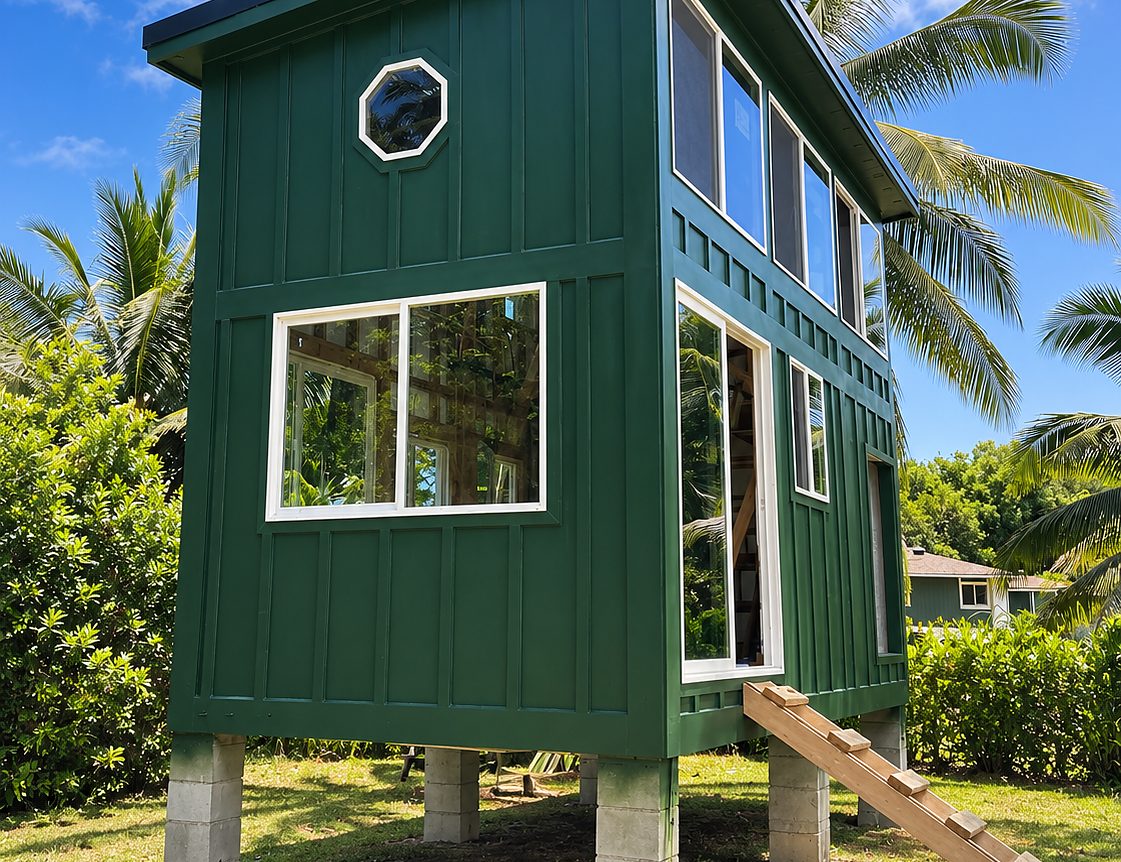Finished green board-and-batten tiny house on concrete piers with a shed roof and palms behind