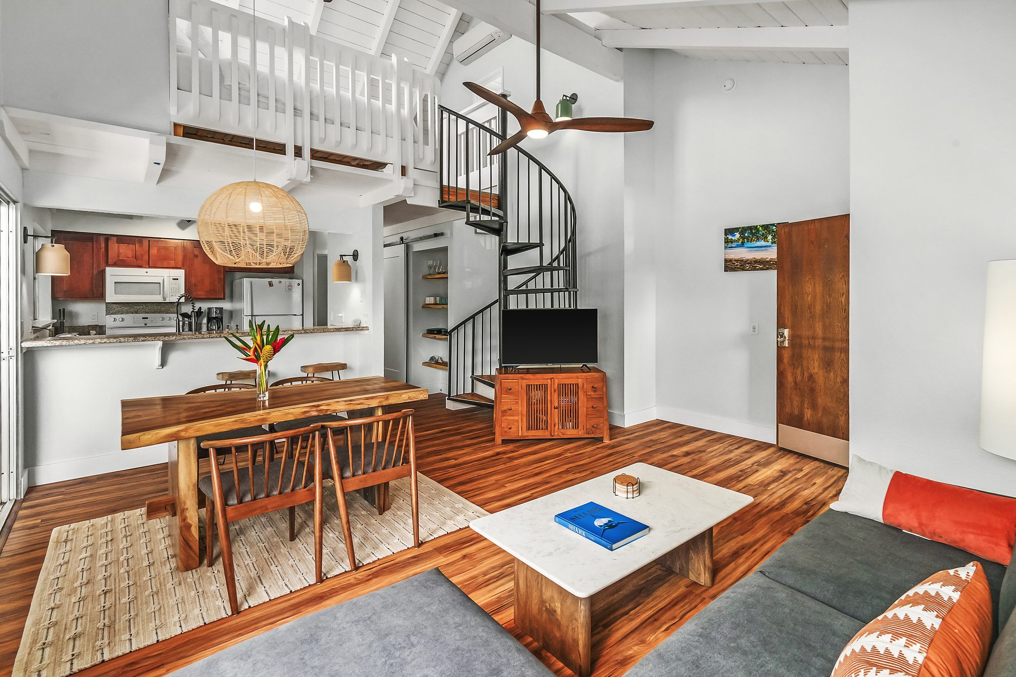 Living room with a vaulted white ceiling, a spiral stair to the loft, a live-edge dining table and the kitchen beyond