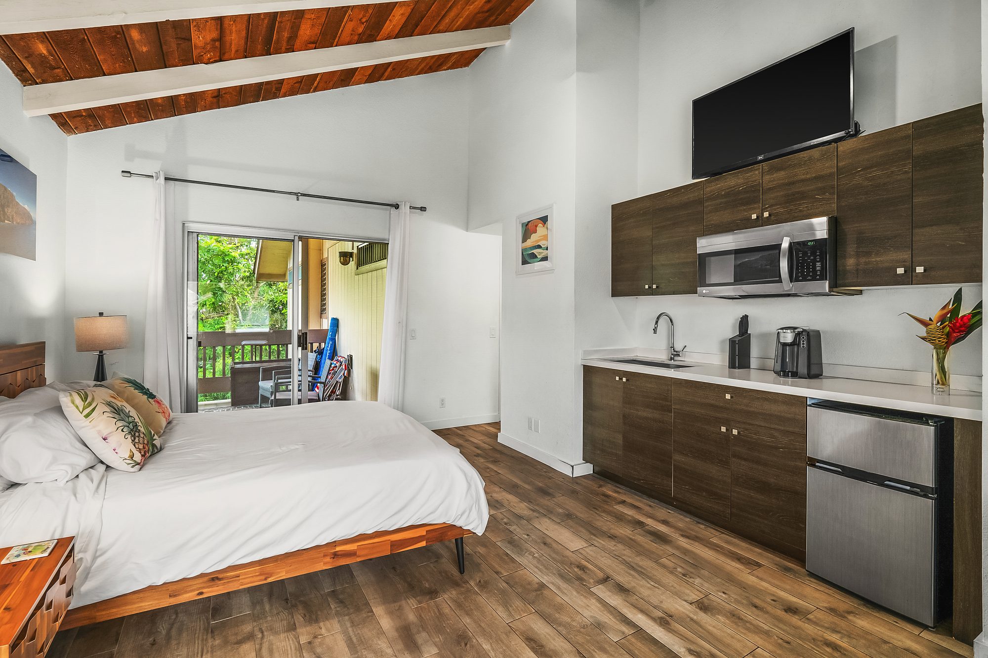 Studio under a wood ceiling with white beams, a bed, kitchenette and window to the trees