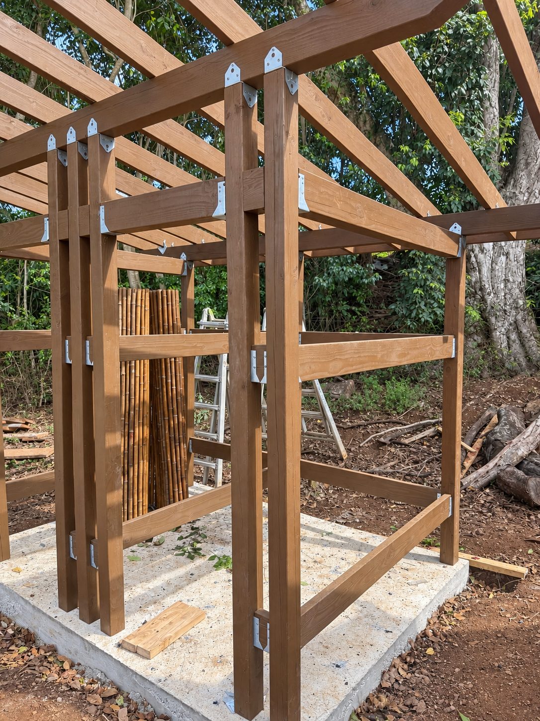 The lumber frame of an outdoor shower on a concrete slab, with hangers and a stack of bamboo poles inside