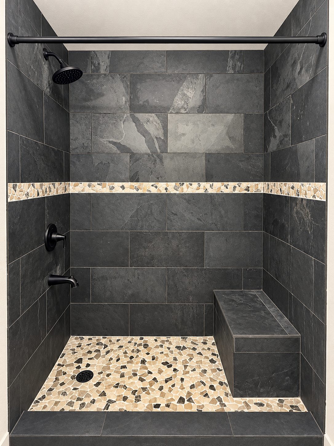 Walk-in shower in dark slate tile with a pebble band, pebble floor and a bench