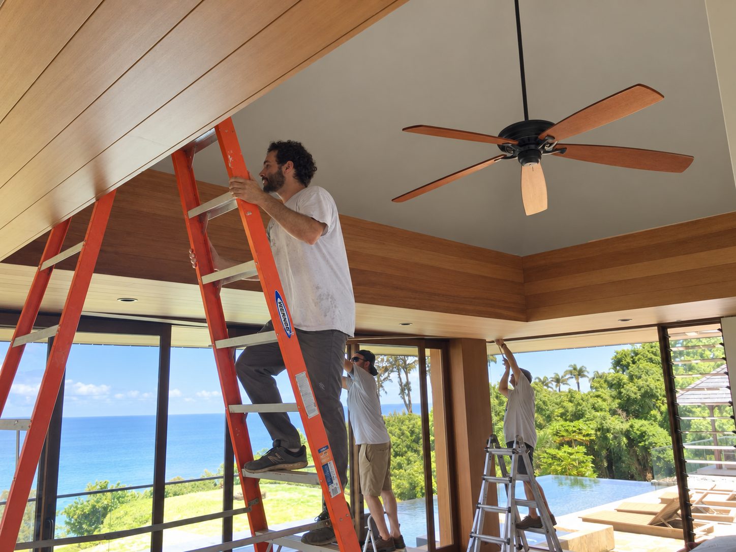 Two carpenters on ladders fitting the wood ceiling in a room open to the ocean and the pool